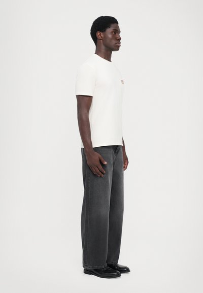 White short-sleeve t-shirt with a small logo on the chest, paired with loose-fitting dark gray jeans and black shoes, viewed from the side.