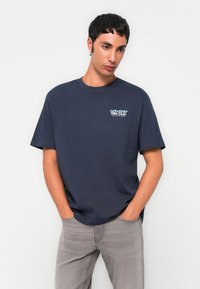 Joven con una camiseta azul marino de "Hike Club" y jeans gris claro, de pie con las manos en los bolsillos frente a un fondo blanco liso.