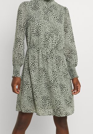 Robe courte en tissu léger vert clair avec des taches noires, dotée de manches longues bouffantes, d'un col montant et d'une taille élastique pour cintrer la silhouette.