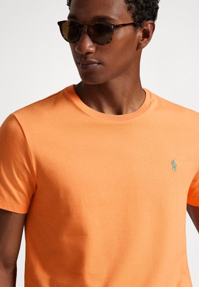 Πορτοκαλί κοντομάνικη μπλούζα από λείο ύφασμα, με μικρό πράσινο λογότυπο πόλο στο στήθος. Ο λαιμός είναι στρογγυλός και ριμπ.
