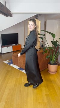 Dlhé čierne šaty s hladkou textúrou, opásaným pásom a voľnou siluetou. Doplnkom sú robustné čierne topánky, všetko situované v jasnom vnútornom priestore.