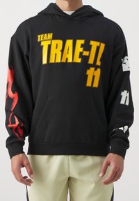 Sweat à capuche noir avec « TEAM TRAE-TL » et « 11 » en caractères gras jaunes. Présente des motifs de flammes rouges sur une manche et des accents blancs sur l'autre.