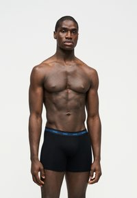 Μυώδης άνδρας με σκουρόχρωμο δέρμα όρθιος, φοράει μαύρα boxer briefs με μπλε ζωνάρι μέσης που γράφει "HUGO", μπροστά σε απλό φωτεινό φόντο.