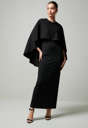 Schwarz Abendkleid mit kapuzenartigem Überwurf, figurbetontem Schnitt und bodenlangem Rock. Kombiniert mit schwarzen Riemchenheels.