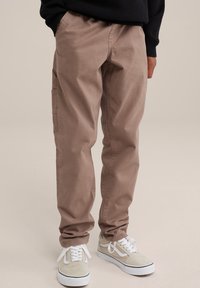 Pantalones cargo de color marrón claro con un ajuste relajado, que presentan bolsillos laterales y una cintura ajustada, combinados con zapatillas beige y blancas.