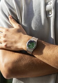 Stainless steel wristwatch with a green dial, silver accents, and a metal band, worn on a tanned arm against a textured gray shirt.