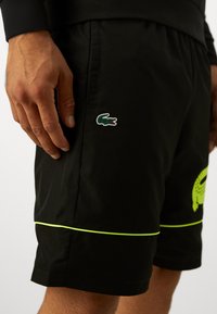 Calções desportivos pretos com detalhes em verde néon, apresentando o logo do crocodilo Lacoste do lado esquerdo. Feitos de material leve, com acabamento texturizado.