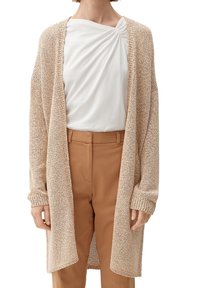 Cardigan beige lavorato a maglia con apertura frontale, maniche lunghe e polsini a coste. Indossato sopra una maglietta bianca e pantaloni color sabbia, con una vestibilità comoda.