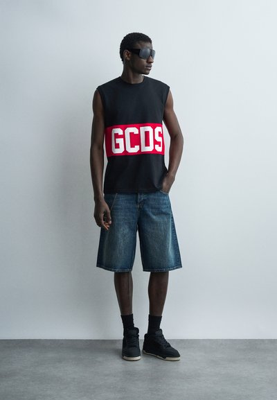 Άντρας που στέκεται, φοράει μαύρο κοντομάνικο μπλουζάκι με κόκκινο λογότυπο GCDS, τζιν σορτς μέχρι το γόνατο, μαύρες κάλτσες, αθλητικά παπούτσια και σκούρα γυαλιά ηλίου.
