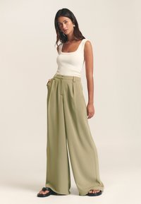 Bela brezrokavnik s kvadratnim izrezom, kombiniran z zelenimi širokimi hlačami z visokim pasom. Gladek material in sproščen kroj. Črne sandale.