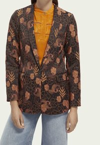 Marynarka z czarnego materiału z pomarańczowymi wzorami kwiatowymi i winorośli, o dopasowanym kroju, z wyłogami i dwiema przednimi kieszeniami. Nałożona na pomarańczowy top.