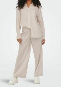 Camisa de lino de color beige claro con botones y bolsillo, combinada con pantalones de lino de pierna ancha. Zapatillas blancas completan el look. Textura suave.