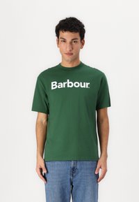 Πράσινο βαμβακερό T-shirt με λευκό λογότυπο "Barbour" μπροστά, κοντά μανίκια και κλασικό στρογγυλό λαιμό, σε συνδυασμό με μπλε τζιν.