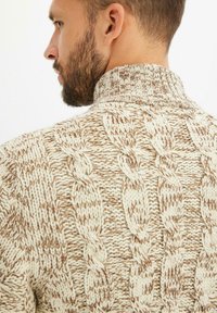 Maglione a treccia in crema e marrone chiaro, con colletto alto e motivo testurizzato. Il tessuto appare morbido e spesso.