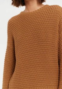 Pull en tricot texturé marron avec un col rond, présentant une coupe ample et des poignets côtelés. Le tissu possède un motif tissé pour plus de détails.