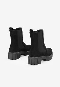 Botas de tobillo de ante negro con paneles laterales elásticos, suelas de goma gruesa y costuras en contraste. Punta redondeada y lengüetas para una fácil puesta.
