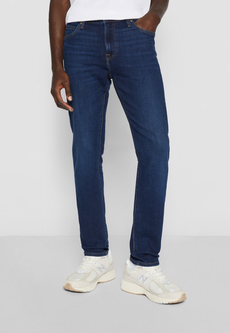 Jeans de mezclilla azul oscuro con un diseño ajustado, que presentan un estilo clásico de cinco bolsillos y costuras en contraste. Llevados con zapatillas blancas.