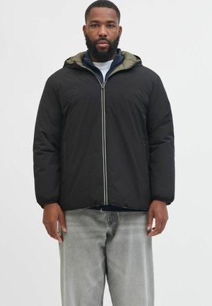 Mann trägt eine schwarze Kapuzenjacke mit Reißverschluss über einer dunkelblauen Innenschicht und hellgrauen Jeans, steht vor einem weißen Hintergrund.