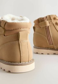Braune Winterstiefel mit weißem Schafslederfutter, Nähdetails, seitlichem Reißverschluss und dicken, off-white Sohlen vor einem neutralen Hintergrund.