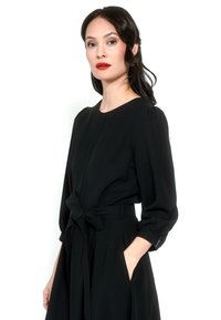 Kobieta z ciemnymi włosami luźno związanymi z tyłu, ubrana w czarną sukienkę z długim rękawem i paskiem w talii, stojąca na białym tle.