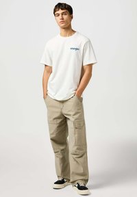 Biała bawełniana koszulka z niebieskim logo "Wrangler", połączona z beżowymi spodniami cargo z kieszeniami i luźnym krojem, uzupełniona czarnymi trampkami.