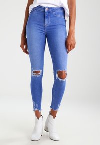 Jeans skinny de mezclilla azul con desgastes en las rodillas y dobladillos deshilachados, combinados con botas blancas hasta el tobillo. Textura suave, diseño ajustado.