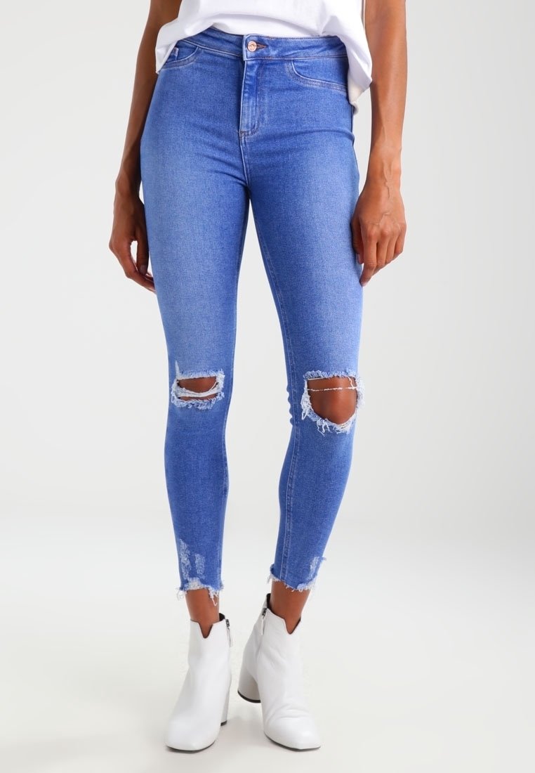 Jeans skinny de mezclilla azul con desgastes en las rodillas y dobladillos deshilachados, combinados con botas blancas hasta el tobillo. Textura suave, diseño ajustado.