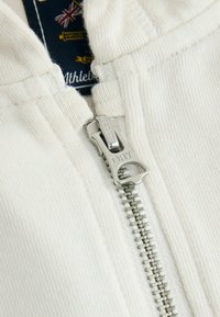 Witte zip-up sweatshirt van textuurstof; voorzien van een zilveren rits en een label met een vintage Brits ontwerp op een marineblauwe achtergrond.