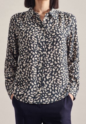 Kvinde iført en sort skjorte med hvide uregelmæssige prikker og mørke bukser, hænderne i lommerne.