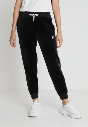Person, die schwarze Nike Velour-Jogginghosen mit weißem Kordelzug und weißen Sneakern trägt, steht vor einem schlichten hellen Hintergrund.