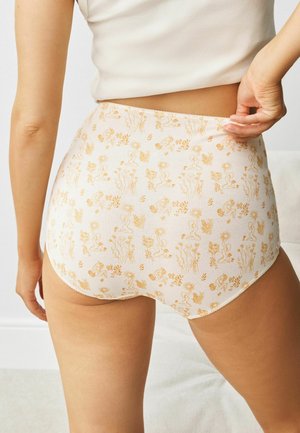 Woman wearing high-waisted, cream underwear with a yellow floral and abstract pattern, adjusting the waistband with her right hand.