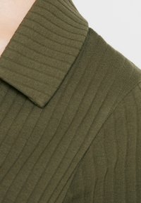 Olivgrüner, gerippter Strickstoff mit einem strukturierten Kragen und einer glatten Textur. Das Design hebt vertikale Streifen und eine taillierte Passform hervor.