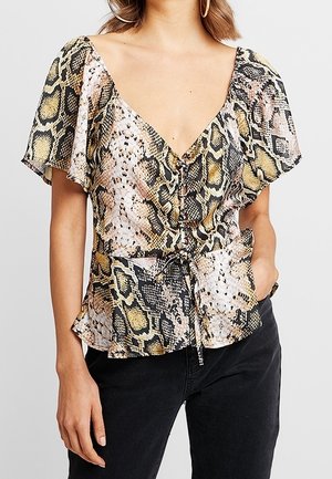 Femme portant un chemisier à manches courtes, col en V avec motif serpent et un pantalon noir, une main dans la poche, sur fond gris clair.