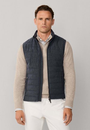 Chaleco acolchado azul marino con cuello alto y cierre de cremallera, superpuesto sobre un suéter beige. El modelo lleva pantalones blancos, mostrando un look casual y a medida.