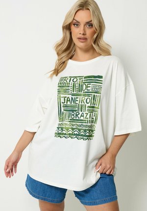 Lyshåret kvinde iført oversized hvid T-shirt med grøn "Rio de Janeiro Brazil" grafik og blå denim shorts, stående mod ensfarvet baggrund.