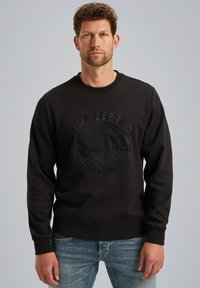 Sudadera negra hecha de un tejido grueso, con un diseño de logotipo en relieve en el centro, cuello redondo acanalado y puños. Llevada con jeans azules.