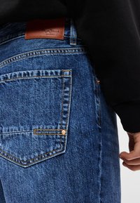 Jeans de mezclilla en azul medio con una textura suave. Presentan costuras en contraste, un parche de cuero marrón claro en la cintura y detalles de herrajes de cobre.