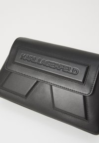 Clutch in pelle nera con superficie testurizzata, accenti geometrici e logo impresso. Chiusura con pattina su un design angolare.