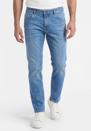 Mann trägt hellblaue, eng anliegende Jeans und weiße Turnschuhe, steht vor einem einfarbigen Hintergrund.