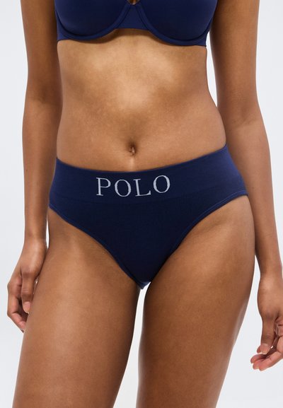 Femme portant des sous-vêtements taille haute bleu marine avec le texte "POLO" sur la ceinture et un soutien-gorge assorti, vue de mi-torse jusqu'aux cuisses.