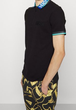 Polo noir à manches courtes avec col bleu Versace et logo subtil "Versace Jeans Couture", associé à un pantalon noir avec imprimé chaîne dorée.