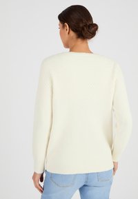Crème-kleurige trui met een gestructureerde, patroonde achterkant; lange mouwen; geribbelde manchetten en zoom; gedragen met lichtblauwe jeans.