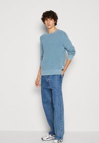 Helles blauer gerippter Pullover mit Rundhalsausschnitt, lockerer Passform und langen Ärmeln, kombiniert mit blauen geraden Jeans und weißen Sneakers.
