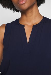 Top senza maniche di colore blu navy con scollatura a V e dettagli di cucitura, realizzato in tessuto liscio. Focus sulla scollatura e sulla zona delle spalle.