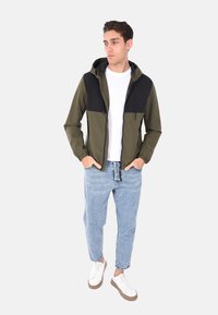 Giacca con cappuccio di colore verde con spalle nere, con zip frontale, polsini elasticizzati, indossata sopra una camicia bianca e jeans in denim azzurro chiaro, abbinata a sneaker bianche.