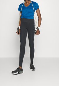 Sportovní outfit zahrnující modré tričko s výstřihem do V, černé těsně přiléhavé legíny a černé tenisky s bílými akcenty, držící posilovací pásy.