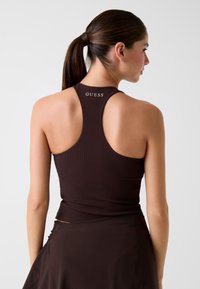 Débardeur marron côtelé avec un dos nageur. Logo "GUESS" imprimé en couleur contrastante sur le haut du dos. Texture lisse.