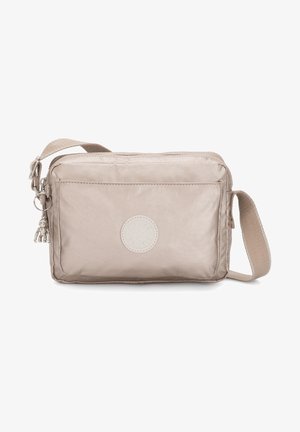 Beżowa torba crossbody wykonana z gładkiego materiału. Posiada przednią kieszeń, okrągłe logo oraz dekoracyjną zawieszkę przymocowaną do zamka.