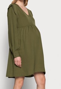 Oliivinvihreä, polvimittainen mekko, jossa on V-pääntie ja rypistetty vyötärö. Pitkät hihansuut napilla ja hienovarainen ribbitekstuuri.