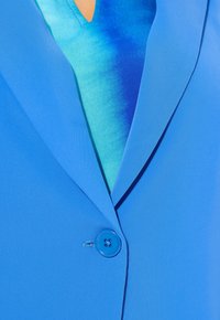 Sininen bleiseri, joka on valmistettu sileästä kankaasta, jossa on yksi nappi, ja sen alla on kerrostettu, liukuväri sinivihreä paita. Yksinkertainen kauluksen muotoilu.